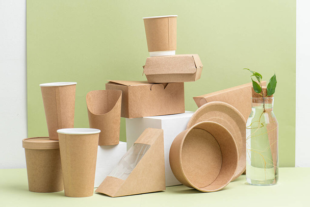 Eco-friendly disposable paper containers for food and drinks over green background. Side view on composition. Some objects placed on white cube pedestals, some placed on side. Plant branch in bottle. - Foto, Imagem