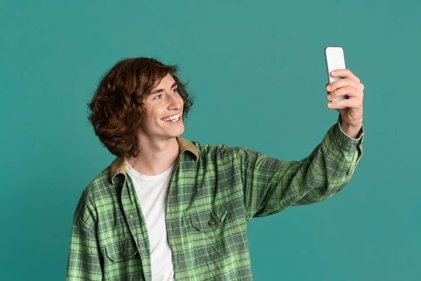 Curly teenage guy with smartphone taking selfie on color background - Фото, изображение