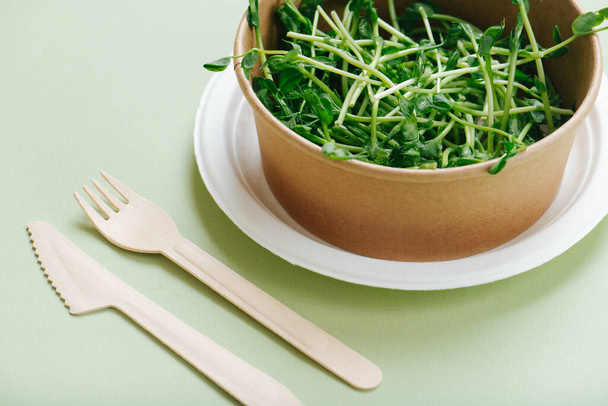 Eco-friendly dinner set. Paper bowl full of pea sprouts and bamboo utensils on the side. Over green background. Disposable and compostable dishware. - Fotoğraf, Görsel