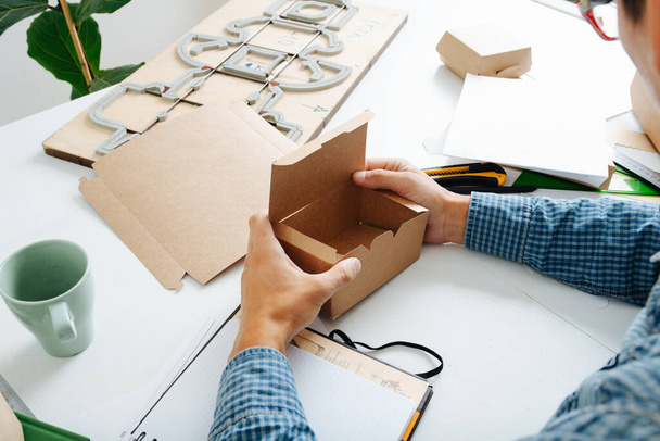 Male box maker working behind a table in a private workshop, folding, shaping a box, tools scattered across. Sheets, boxes and special board with a cutting form. - 写真・画像