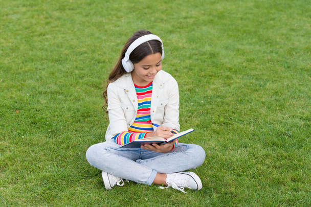 write song lyrics. happy childhood concept. kid write memory in diary. schoolgirl relax with notebook. happy child listen music in headphones. small girl on green grass. kid enjoy song in park nature - Foto, imagen