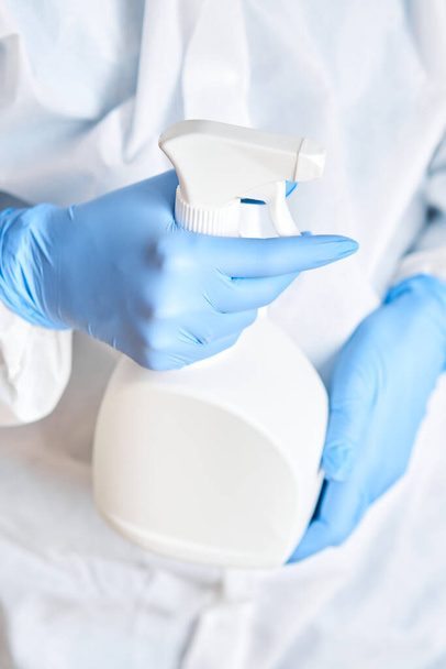 Closeup of a white spray bottle in woman hands in a protective suit and blue gloves. Disinfection, cleaning and washing. COVID-19. Prevention of coronavirus infection. - Fotó, kép