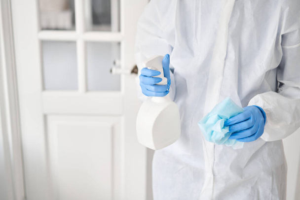 Koruyucu takım elbiseli ve mavi eldivenli bir kadının elindeki beyaz sprey şişenin yakın çekimi. Dezenfeksiyon, temizlik ve yıkama. COVID-19. Koronavirüs enfeksiyonunun önlenmesi. - Fotoğraf, Görsel