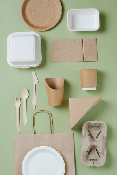 Eco-friendly disposable dishware and bamboo utensils over green background. Paper and cornstarch cases, cups, packets and dishes. Top view. - Foto, Imagen