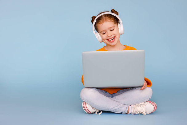 Cuerpo completo niña positiva en los auriculares sonriendo y viendo dibujos animados en el ordenador portátil moderno mientras está sentado con las piernas cruzadas en el fondo azul
 - Foto, Imagen