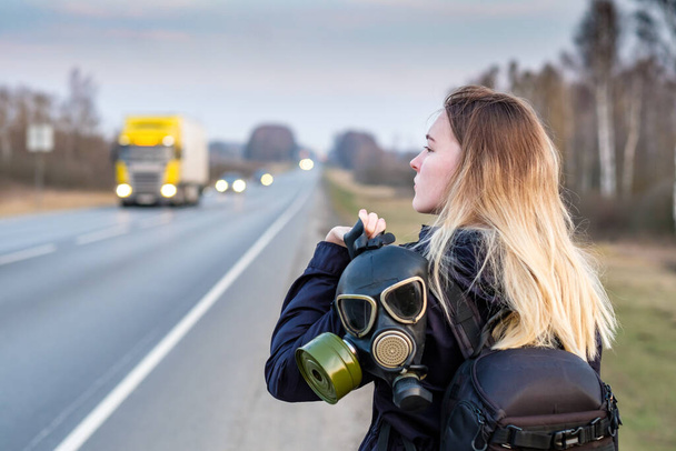 A girl with a black gas mask on his shoulder stands on the edge of a suburban highway. The girl is trying to stop passing cars in order to leave the city in which the epidemic of coronavirus. - 写真・画像
