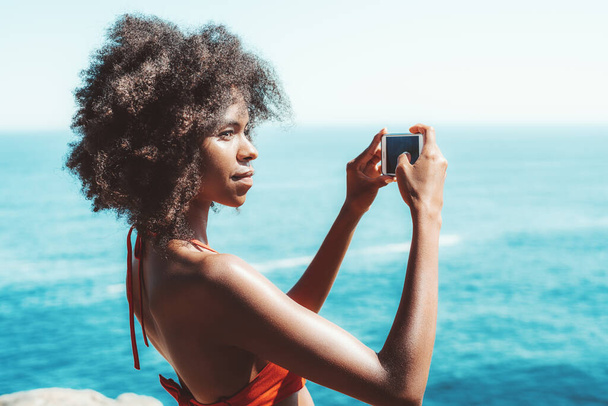 Portrait of a dazzling African-American girl shooting a seascape on her smartphone; young charming Brazilian female with a curly Afro hair and in swimwear is photographing resort using the cellphone - Foto, Imagem