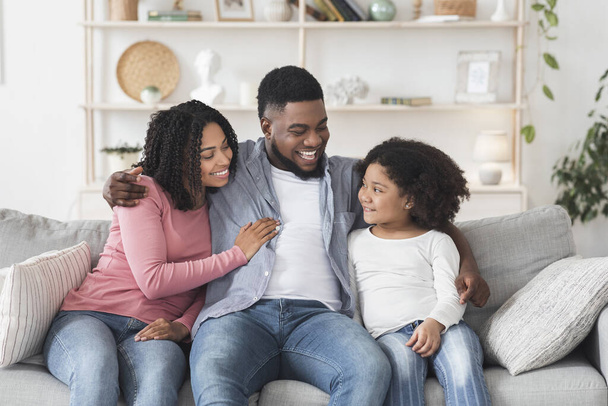 Любящий чёрный мужчина обнимает жену и дочь на диване дома
 - Фото, изображение