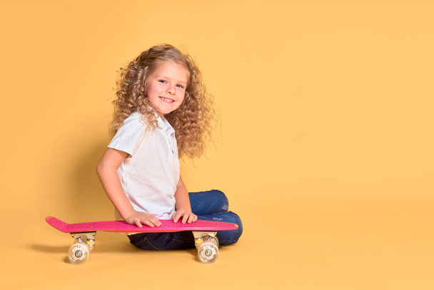 Active and happy girl with curly hair, headphones having fun with penny board, smiling face stand skateboard. Penny board cute skateboard for girls. Lets ride. Girl with penny board yellow background - Fotografie, Obrázek