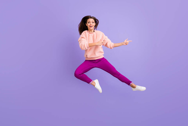 Full length photo of cheerful lady jumping high indicating fingers empty space amazing sale wear casual fluffy pullover pants shoes isolated purple color background - Fotó, kép