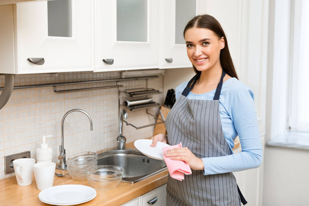 Νεαρή γυναίκα στεγνώνει τα πιάτα, τα σκουπίζει. - Φωτογραφία, εικόνα