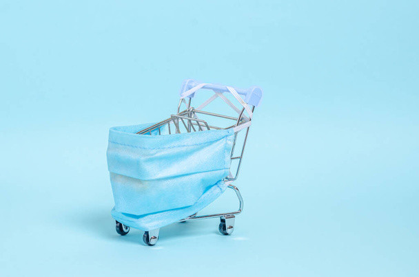 Decorative small shopping trolley with protective sterile medical mask on a blue background. - Foto, imagen