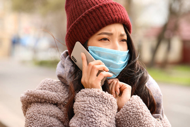 Nuori aasialainen nainen suojelevassa naamiossa puhuu matkapuhelimella kaupungin kadulla. Epidemian käsite
 - Valokuva, kuva