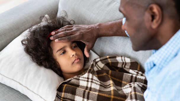 Epidemia wirusa. Starszy Afroamerykanin dotyka swoich wnuczek na czole, sprawdza gorączkę w domu - Zdjęcie, obraz