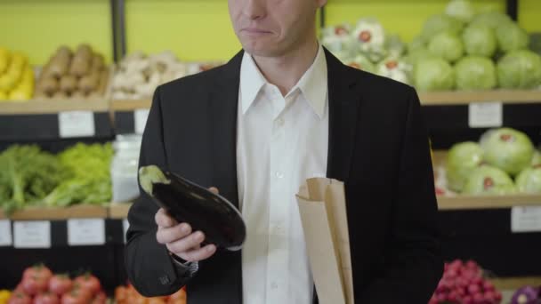 Serious Caucasian man in suit smelling eggplant, putting it into paper pack and leaving. Young businessman purchasing vegetarian food in supermarket. Healthy lifestyle, vegetarianism, consumerism. - Metraje, vídeo