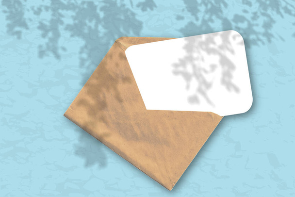 Un sobre con una hoja de papel blanco texturizado sobre el fondo azul de la pared. Mockup superpuesto con las sombras de la planta. La luz natural proyecta sombras desde el follaje del árbol
. - Foto, Imagen