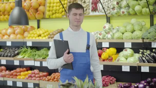Young positive Caucasian employee posing in grocery with folder and eggplant. Portrait of smiling man in uniform working in supermarket. Business, commerce, profession, lifestyle. - Séquence, vidéo