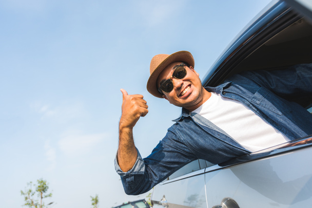 Een reiziger met een hoed op rijdt in een auto en trekt zich dan uit het autoraam. En duimen omhoog Deze foto gaat over een veilige reis met de auto. - Foto, afbeelding