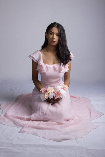 Beautiful young South Asian woman kneeling on the floor in a pink dress and holding a bunch of carnation flowers - 写真・画像
