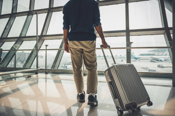 灰色の帽子をかぶった男性旅行者旅行の準備彼はトロリーバッグを持っています彼は世界中を旅行するための大きな空港に立っています. - 写真・画像