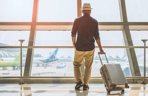 een mannelijke reiziger draagt een grijze hoed hij bereidt zich voor om te reizen hij heeft een trolley tas hij staat op een groot vliegveld voor reizen over de hele wereld. - Foto, afbeelding