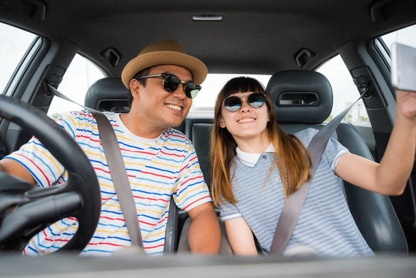 アジア系の男性と女性のカップルが車に座ってスマートフォンを見ています。旅行のコンセプト. - 写真・画像