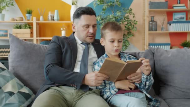 Father in formal clothing reading book to cheerful little boy in apartment - Záběry, video