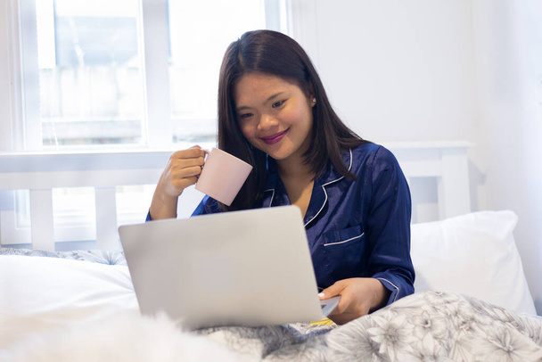 Young asian smiling woman working on laptop while holding a pink mug. Happy morning - Foto, Imagem
