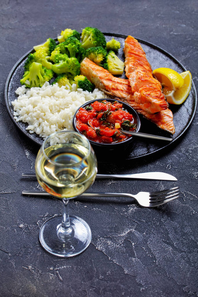 Grilled salmon steak with vegetables: broccoli, tomato, and spinach sauce and long grain rice, lemon wedges on a black platter on a dark concrete table, vertical view - Foto, Imagen