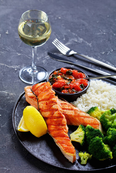 Restaurant serving: salmon steak with vegetables: broccoli, tomato, and spinach sauce and long grain rice, lemon wedges on a black platter with white wine on a dark concrete background with cutlery - Photo, image
