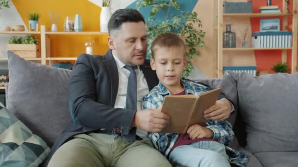 Dad businessman teaching little son to read holding book sitting on couch at home - Filmmaterial, Video