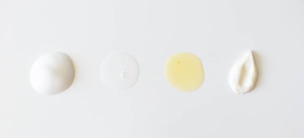 Sample beauty products for facial skin care on white background, above. Cleanser foam, tonic, oil serum essence drop and moisturizer cream smear - 写真・画像