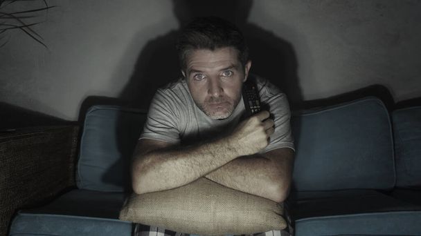 young attractive concentrated and scared man holding TV remote watching horror movie or suspense thriller at night in home couch enjoying film emotion in domestic entertainment concept - Foto, imagen