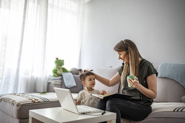 Smiling mom working at home with her child on the sofa while writing an email. Young woman working from home, while in quarantine isolation during the Covid-19 health crisis - Φωτογραφία, εικόνα