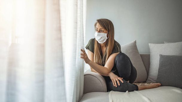 Sad lonely girl isolated stay at home in protective sterile medical mask on face looking at window, bored woman because of Chinese pandemic coronavirus virus covid-19. Quarantine, prevent infection - Zdjęcie, obraz