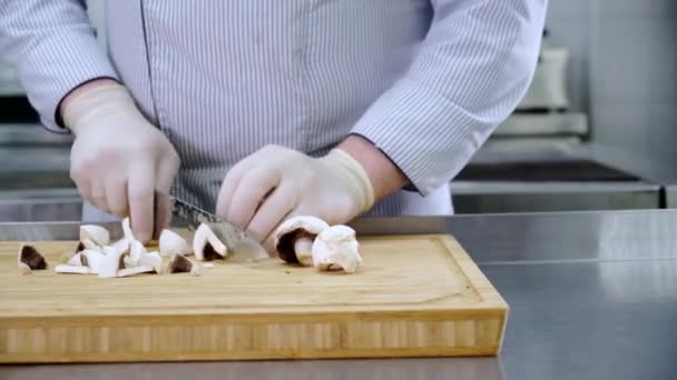 Chef of caucasian man in kitchen of restaurant in gloves cuts raw mushrooms with knife on bamboo wooden cutting board and puts them to side. Dolly slide video - Footage, Video