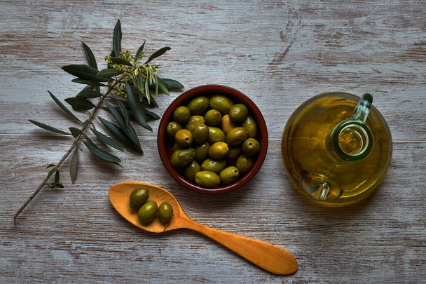 Schaal met groene olijven, houten lepel, olijftak en olie - Foto, afbeelding