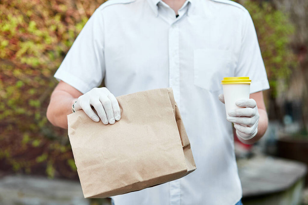 Courier, delivery man in protective mask and medical gloves delivers takeaway food and coffee. Delivery service under quarantine, disease outbreak, coronavirus covid-19 pandemic conditions. Stay home, safe concept. - Foto, imagen