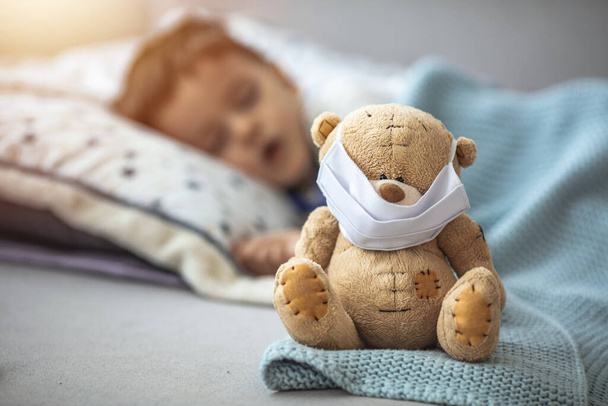 Young child sleeping with teddy bear who is wearing a respiratory mask as a prevention against the Coronavirus Covid-19. Child in home quarantine. Children and illness disease concept - Foto, Bild