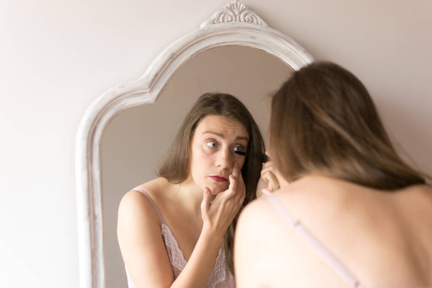 Retrato de mujer joven maquillándose delante de un espejo con herramientas de maquillaje sobre la mesa. Luz natural y planta
. - Foto, imagen