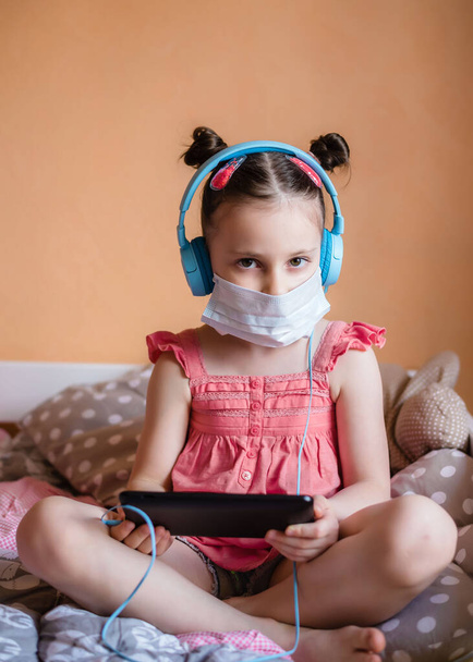 Kid girl wearing protection face mask against coronavirus. Kid in mask at home with tablet/planshet in her hands - Valokuva, kuva