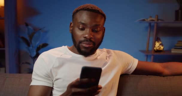 Front view of handsome african man looking at smartphone screen while sitting on couch. Bearded young guy chatting in social networks while spending free time at home. - Кадры, видео