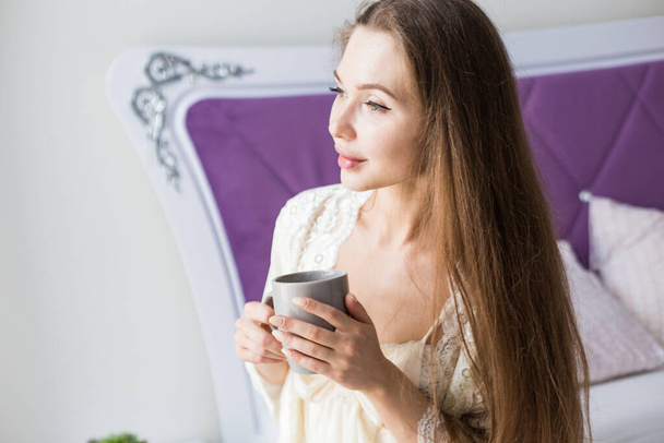 Attractive young girl in nightwear sits on the edge of the bed by the window with a cup of coffee - Fotó, kép