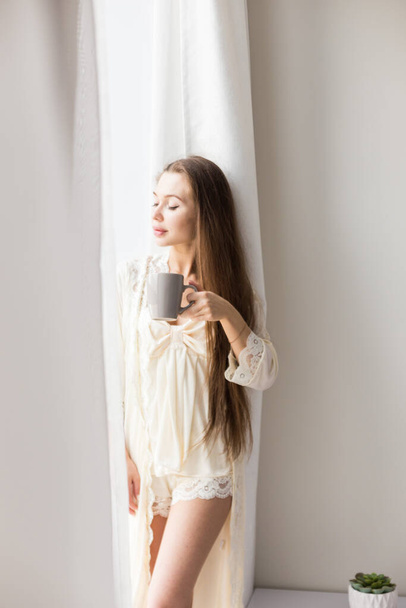 Attractive young girl in night clothes standing by the window with a mug of coffee - Φωτογραφία, εικόνα