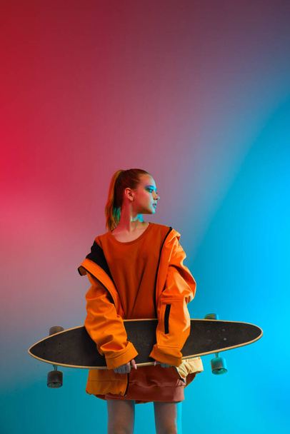 A young girl with bright makeup holds a longboard in her hands on a fashionable neon color background. Portrait of a girl skater - Фото, изображение