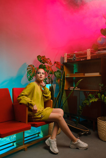 A young attractive girl with bright makeup and in colored clothes sits on a red armchair against a fashionable neon background. - Foto, afbeelding