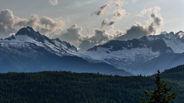 Dağlık bir ormanın karşısındaki karlı dağ tepelerinin görüntüsü ilginç bir gökyüzüne doğru ilerliyor. Dağlar Whistler 'a yakın, British Columbia, Kanada - Fotoğraf, Görsel