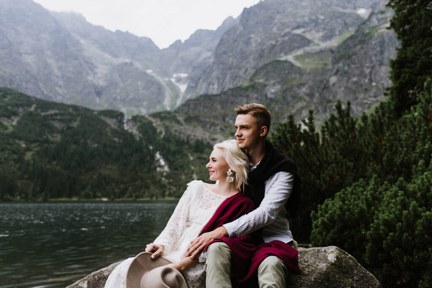 Beautiful bride in a boho dress and groom hug and sit on stones in the mountains near the lake. The bride and groom hug and smile in the mountains. - Photo, Image