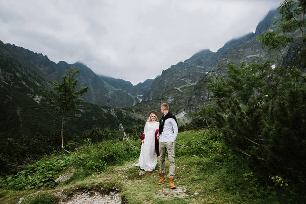 Μια όμορφη νύφη με ένα φόρεμα boho, ένα καπέλο και ένας γαμπρός χαμογελούν και γελάνε στα βουνά. Η νύφη και ο γαμπρός αγκαλιάζονται και περπατούν στα βουνά. - Φωτογραφία, εικόνα