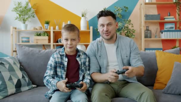 Portrait of father and child playing video game at home, pressing buttons on joystick talking - Metraje, vídeo